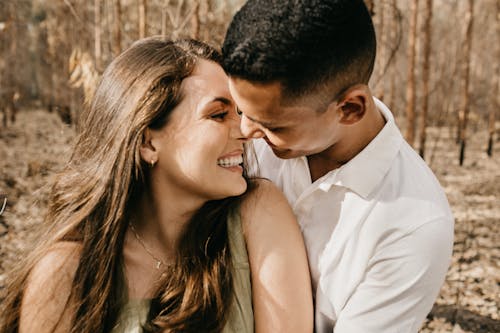 Free Cheerful multiethnic couple embracing gently and smiling happily standing close in forest with bare trees Stock Photo