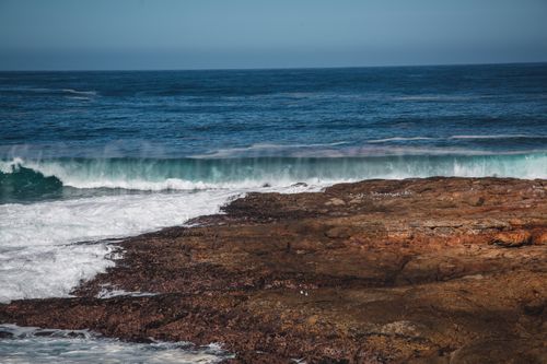 Free Vivid ocean waves crashing onto a rocky shore under a clear blue sky, showcasing nature's raw beauty. Stock Photo