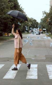 Young woman crossing road with umbrella, surrounded by falling banknotes.