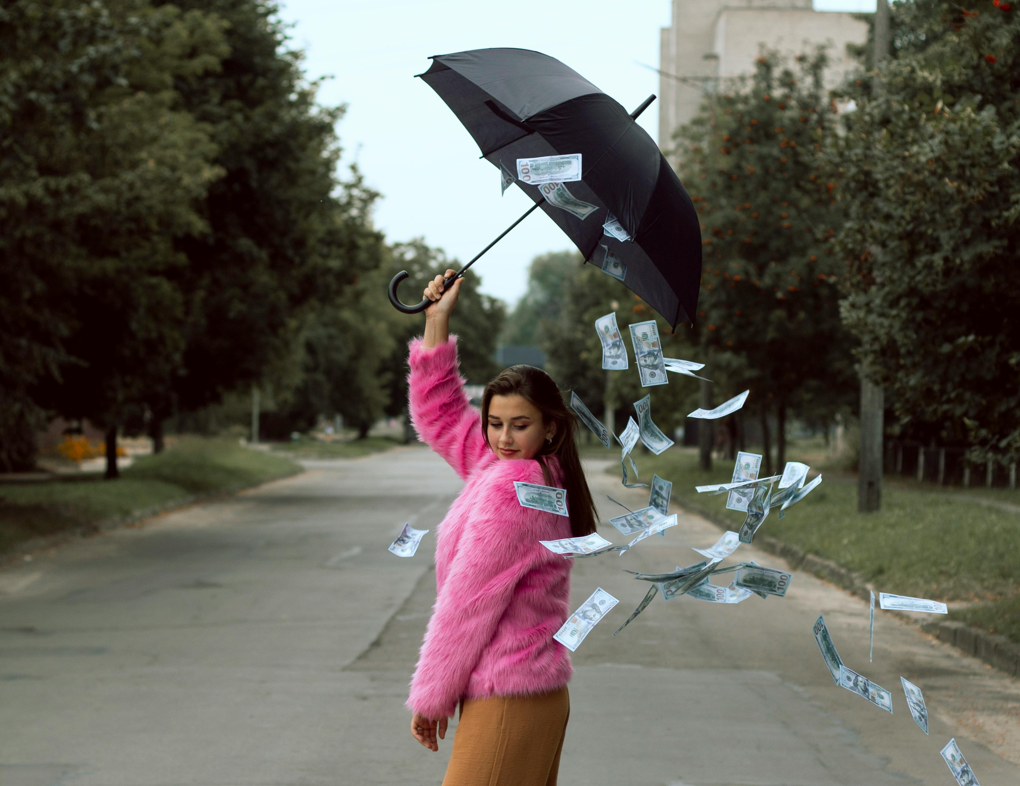 Woman in a vibrant pink fur coat holding an umbrella with cash falling around her, set in a serene outdoor environment.