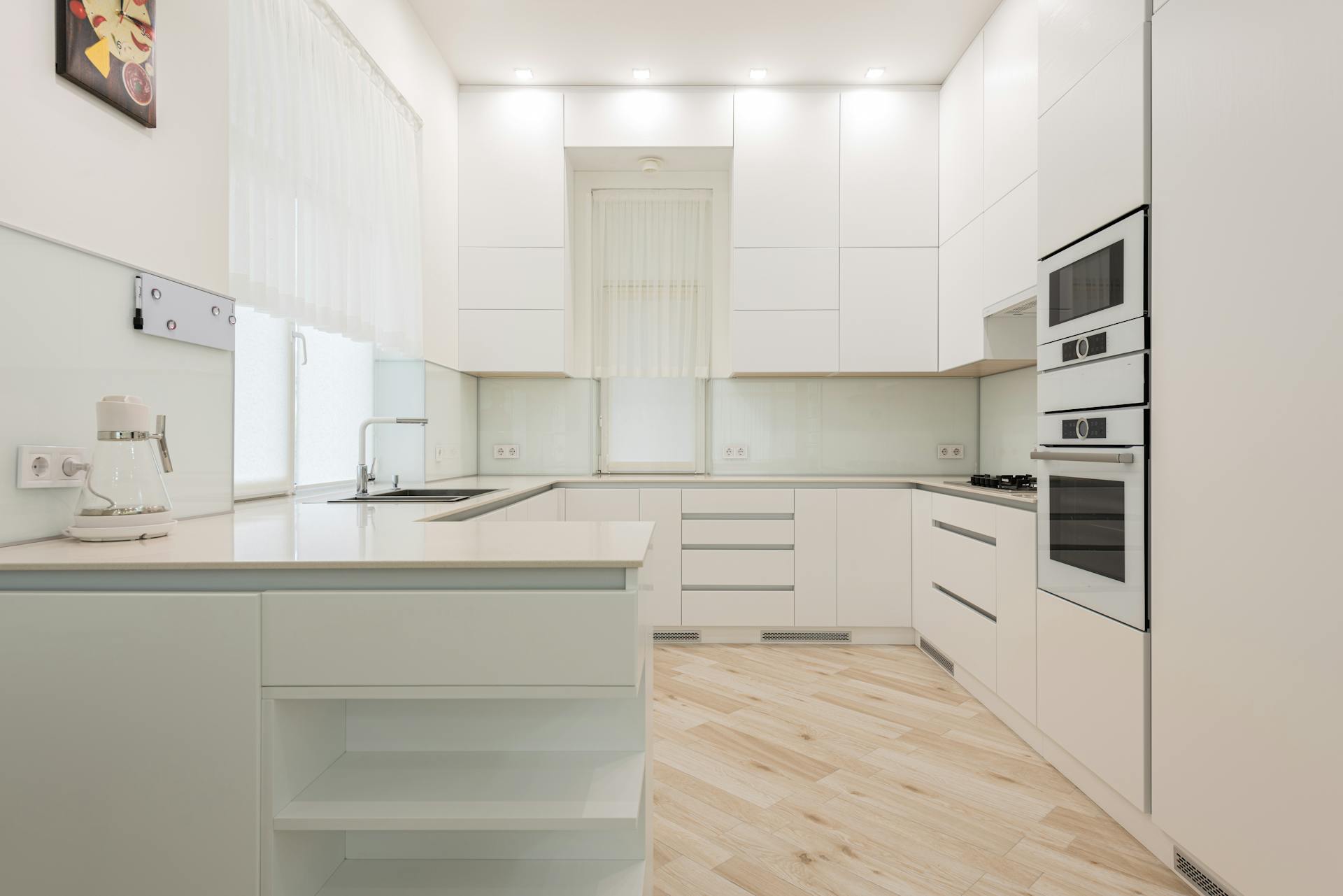 Interior of light kitchen with white cabinets and kettle near sink placed against trendy oven and modern appliances in apartment