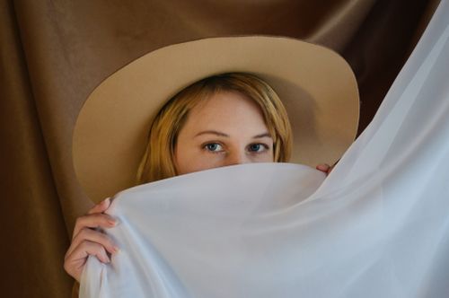 Free Close-up portrait of a woman peeking from behind a curtain with a wide-brimmed hat. Stock Photo