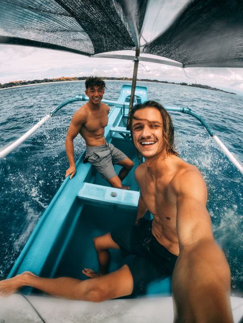 From above of cheerful young shirtless male traveler smiling and taking selfie while floating on boat in wavy ocean with Asian friend