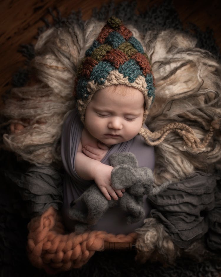 A Cute Baby With Knitted Cap Wrapped With Gray Fabric Holding An Elephant Plush Toy