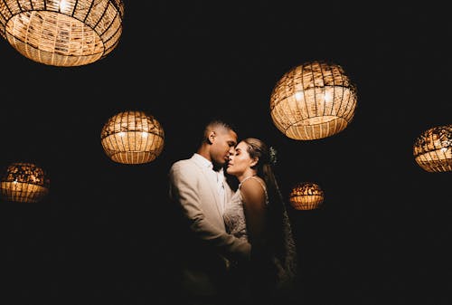 Romantic ethnic newlywed couple wearing classy wedding outfits embracing gently with eyes closed in dark studio decorated with shiny creative lanterns