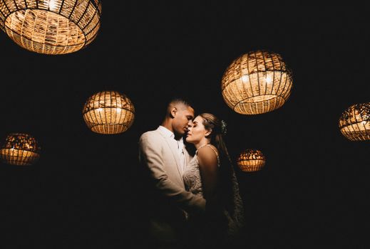 Romantic ethnic newlywed couple wearing classy wedding outfits embracing gently with eyes closed in dark studio decorated with shiny creative lanterns