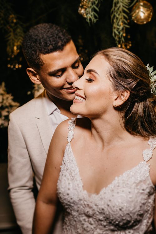 Free Smiling ethnic groom standing with eyes closed behind and hugging cheerful gorgeous bride in posh white dress during wedding celebration Stock Photo