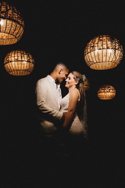 Content young ethnic newlywed couple wearing fancy wedding outfits embracing gently in dark wedding studio decorated with luminous lanterns