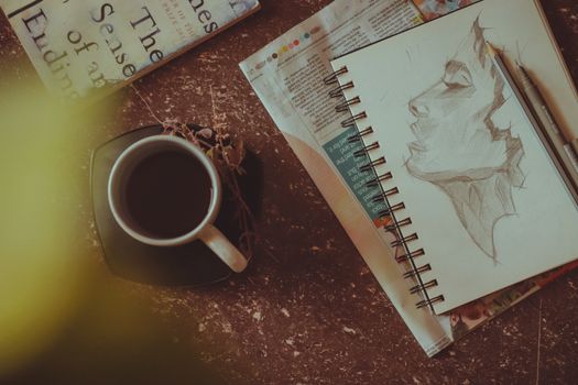 From above of sketchbook with person portrait near pen and pencil next to mug with drink on saucer with herbs on table near book and newspaper in light room