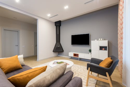 Interior of spacious room with comfortable couch with cushions and armchair placed against wall with TV and white cupboards in apartment with fireplace