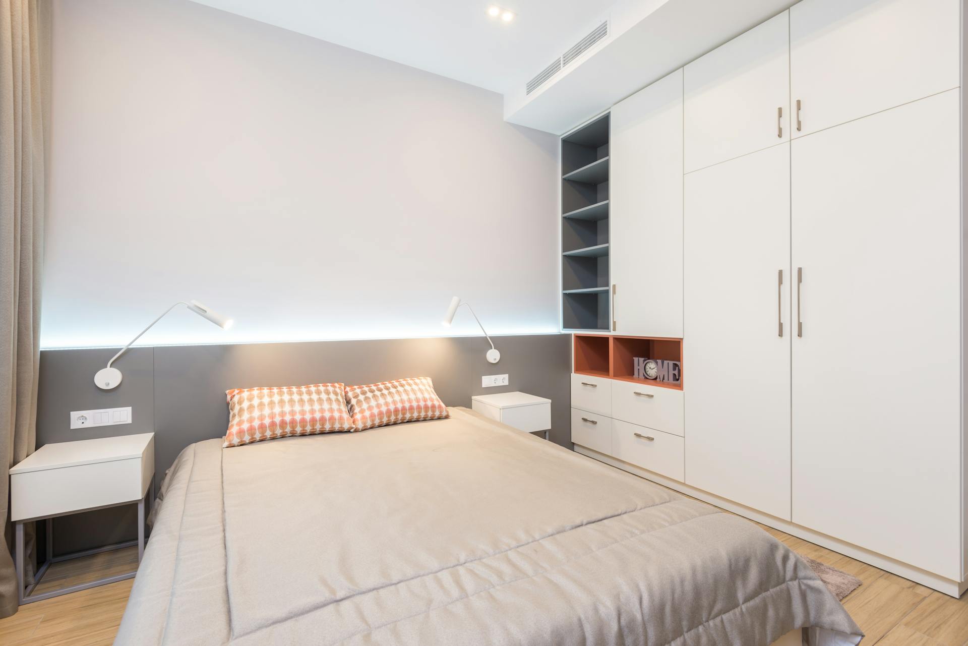 Comfortable bed with pillows placed near bedside tables and white wardrobe in modern spacious bedroom with glowing lamps in apartment