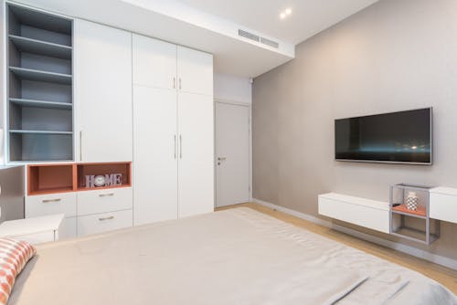 Modern TV set hanging on wall in front of comfortable bed in cozy bedroom with white furniture