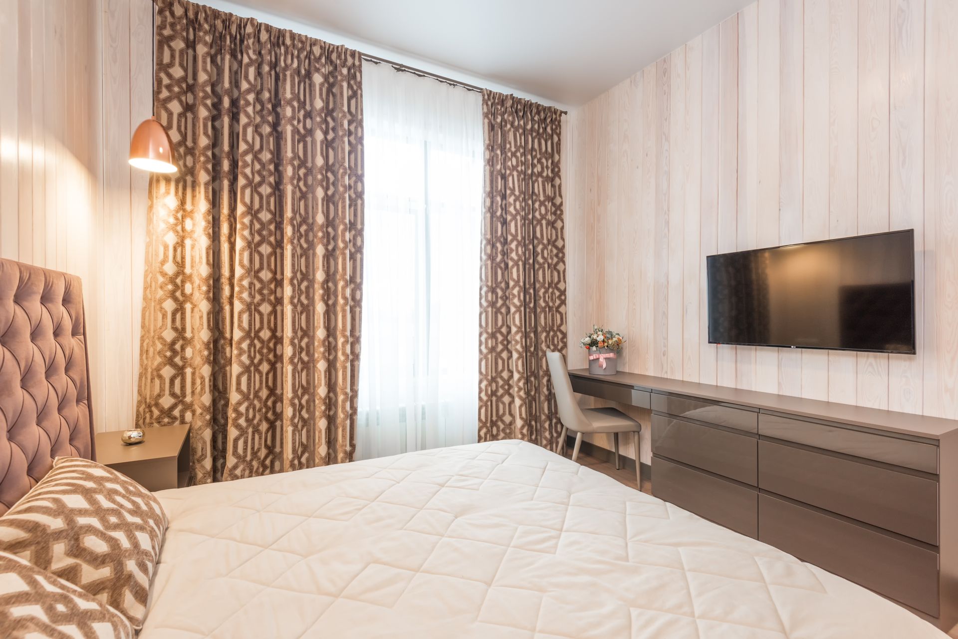 Comfortable bed with soft cushions and headboard in classic styled bedroom with modern TV set and chair