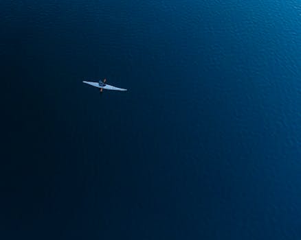 A lone kayaker paddles across a calm blue lake, captured from an aerial perspective.