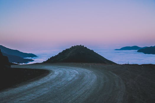 A scenic mountain road curves into the distance under a vibrant twilight sky with fog covering the valleys.