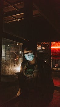 A person with a face mask using a phone at night under low light in an urban environment.