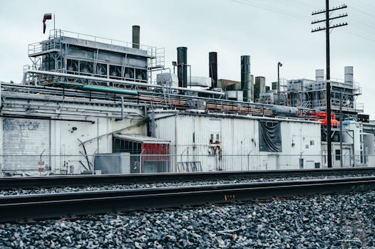A weathered factory building with visible pipelines and railway tracks under an overcast sky.
