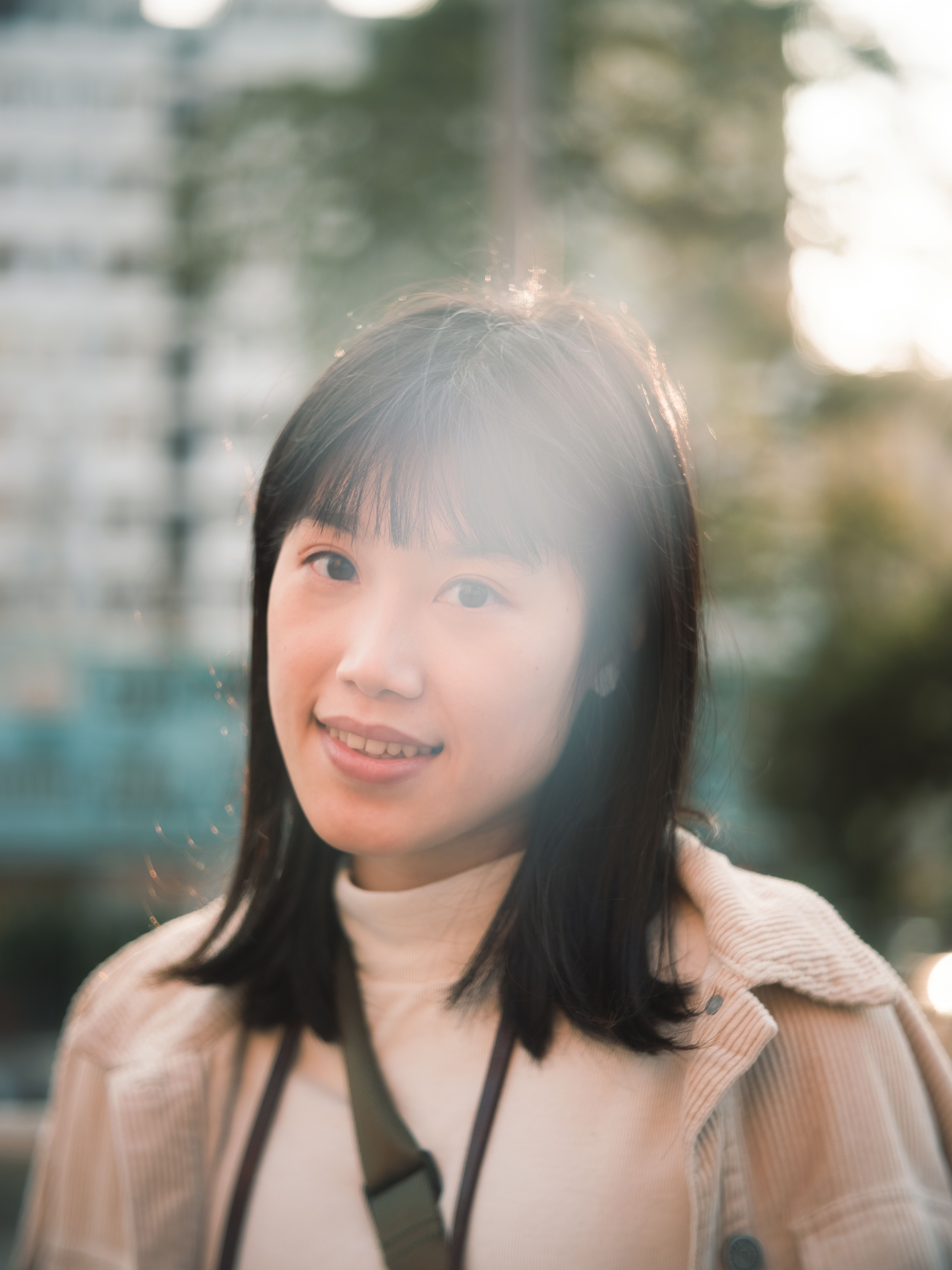 A young woman smiles warmly with sunlight filtering through trees in an urban setting.