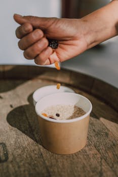 Close-up of a hand adding dried fruits to oatmeal in a container.