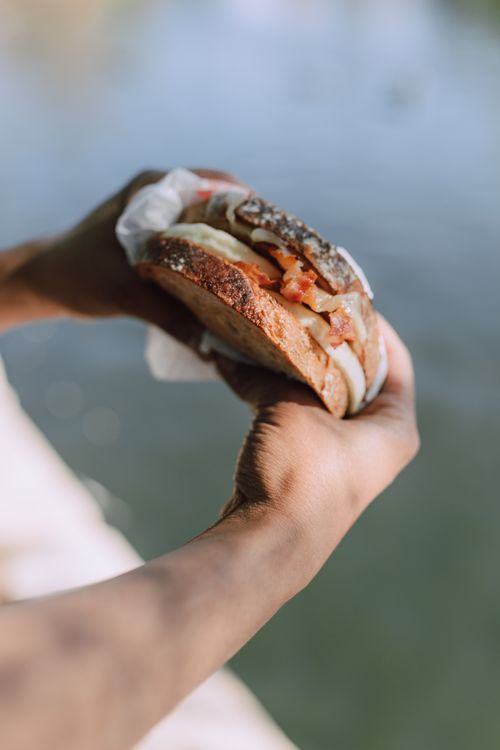 Free A delicious sandwich with bacon, egg, and filling being held by a person near water. Stock Photo