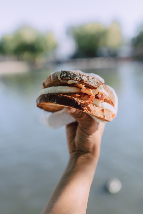 Free A close-up of a hand holding a bacon and egg sandwich against a blurred outdoor background. Stock Photo