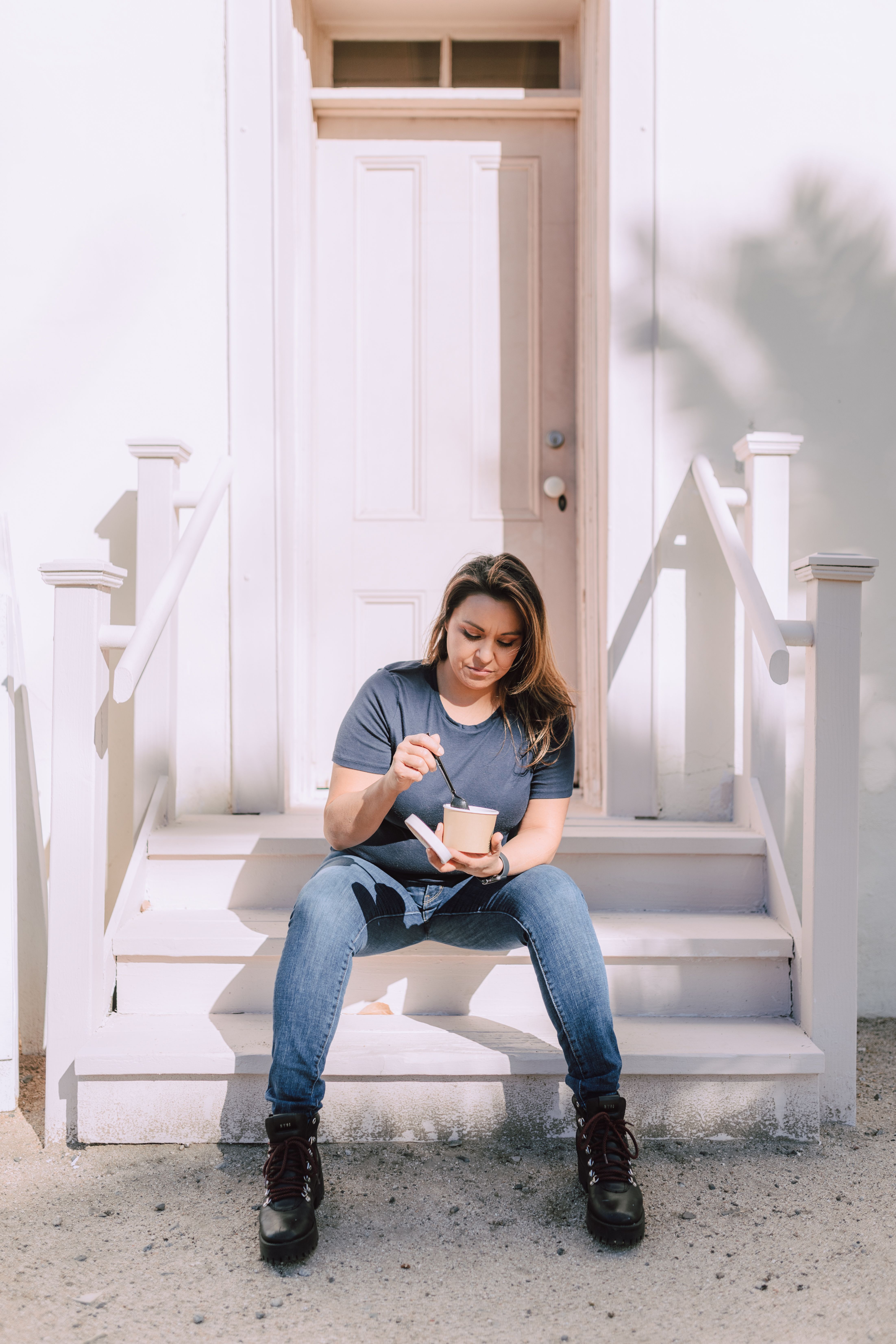 A woman in casual attire eating a takeaway meal while sitting on a sunny doorway step.