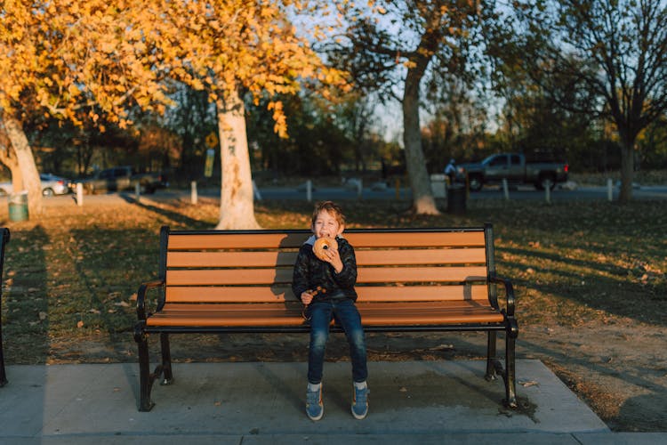 Kid Sitting On A Wooden Bench Eating  A Bagel 