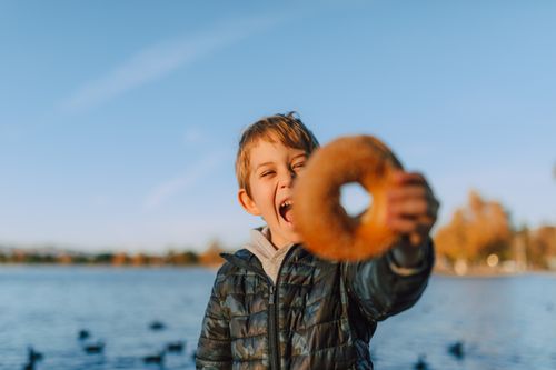 Free Happy child holding a bagel at a lakeside, enjoying a sunny outdoor snack. Stock Photo