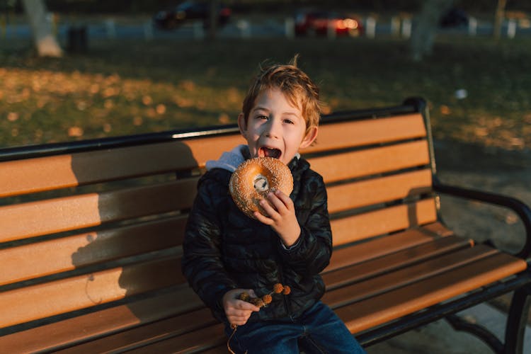 Kid Sitting On A Wooden Bench Eating  A Bagel 