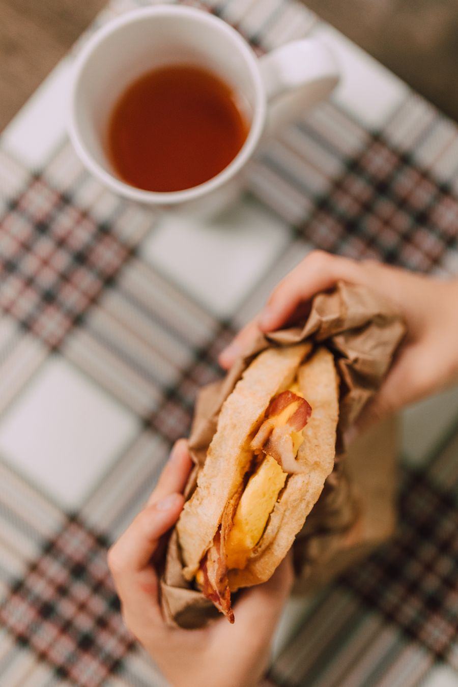 A breakfast sandwich held above a table beside a cup of tea
