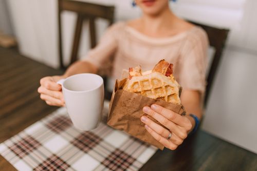 Free A woman holding a waffle with bacon and a cup of coffee, sitting at a table. Stock Photo