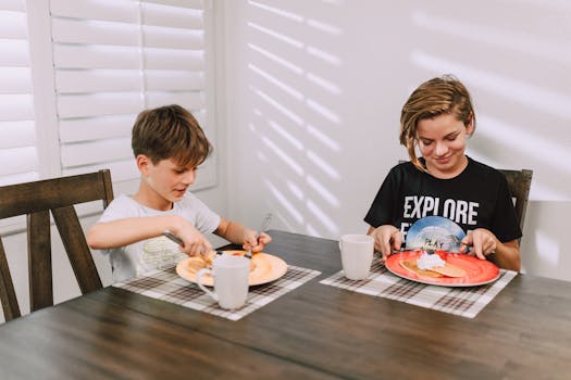 Two boys happily eating breakfast at a wooden table inside a bright room.