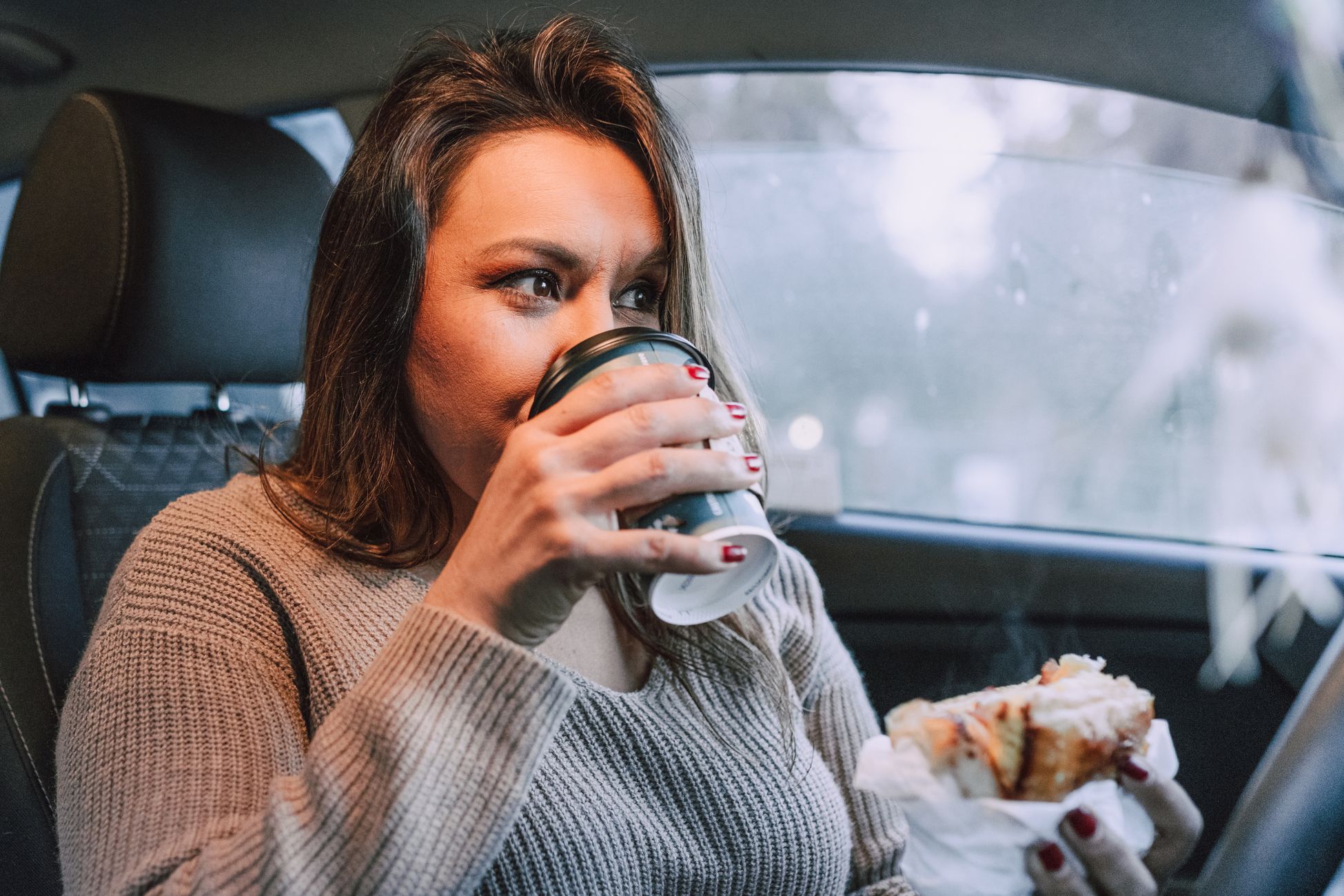 Woman enjoying coffee and pastry inside a car, reflecting a cozy morning routine.