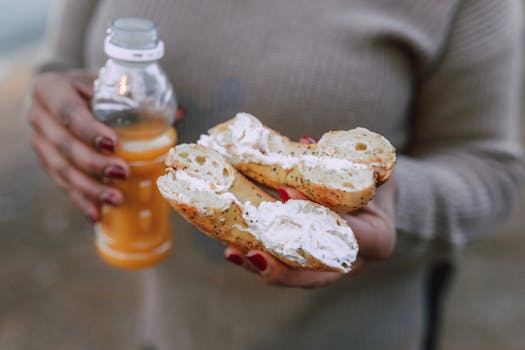 Close-up of hands holding a bagel with cream cheese and a bottle of orange juice outdoors.