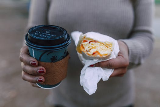 Close-up of a woman holding a hot coffee cup and a breakfast sandwich outdoors.