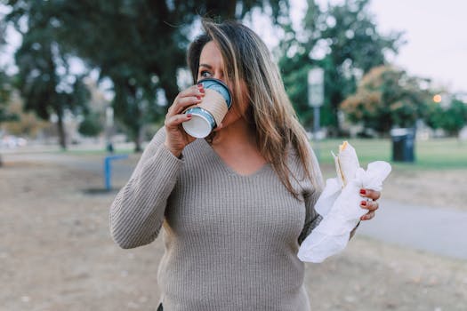 Woman sipping coffee and holding a sandwich in a peaceful park setting.