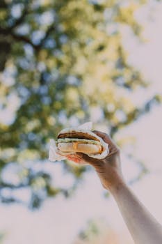 Close-up of a hand holding a burger against a blurred outdoor background.