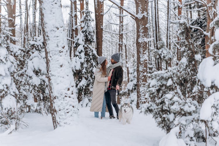 Couple In Winter Clothes Standing In A Snow Covered Forest With Their Dog