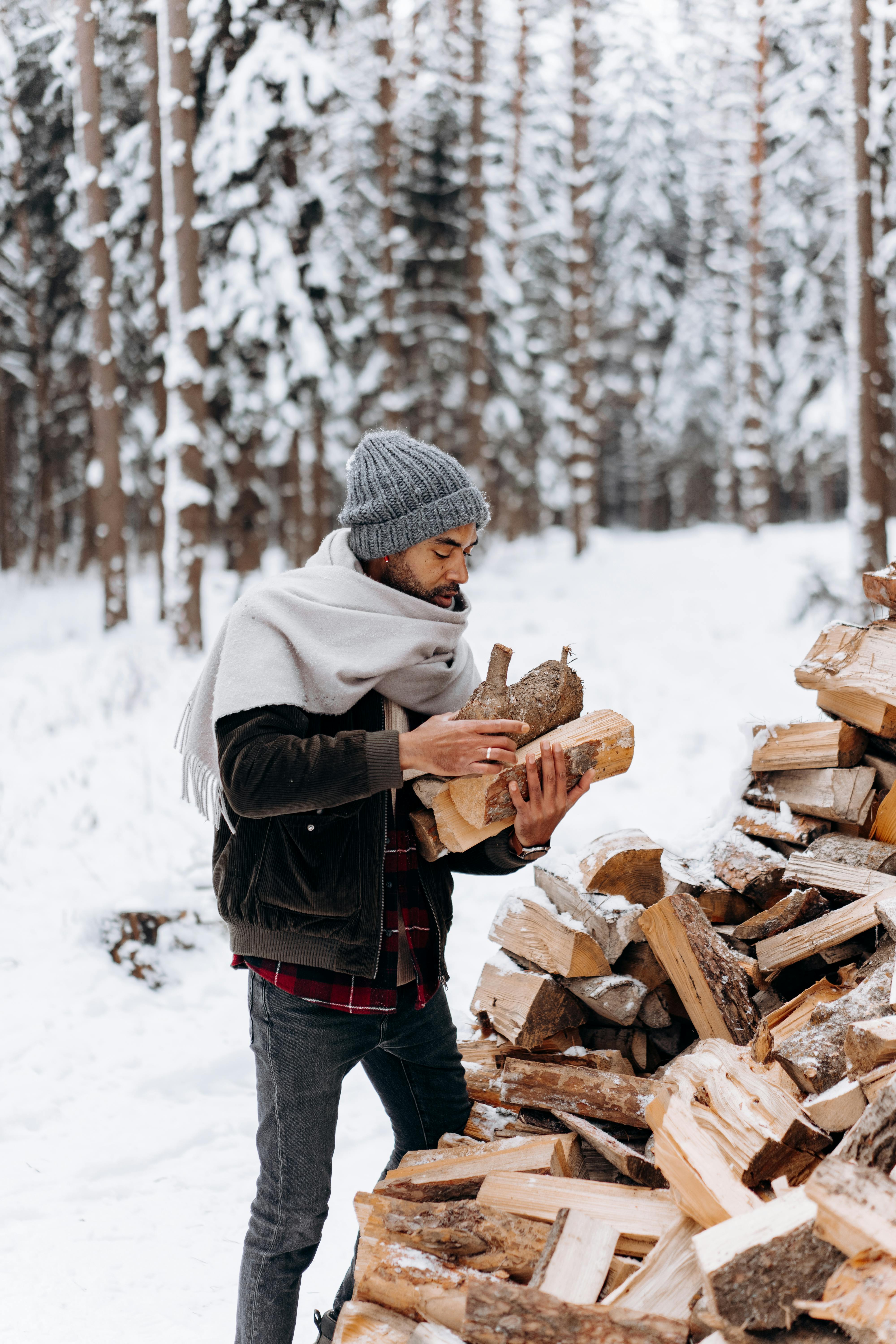 Person gathering logs in snowy winter forest, dressed in beanie and scarf, amidst tall trees.