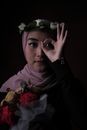Graceful young Muslim female in hijab and floral headband showing OK gesture near eye while standing in dark studio with bunch of artificial flower and looking at camera
