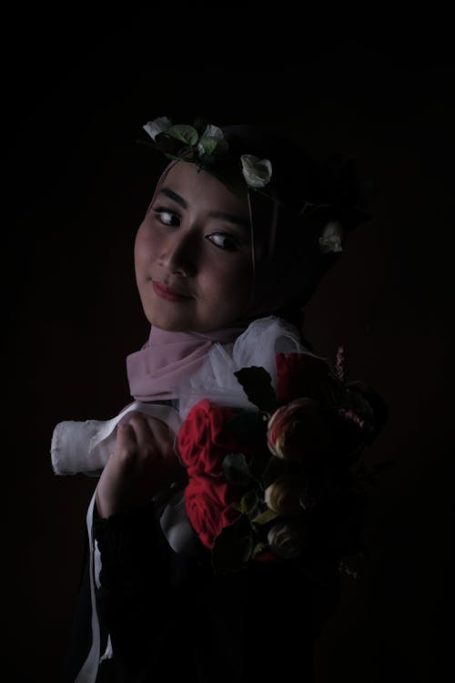Free Side view of positive young Asian lady in headscarf and floral headband smiling and looking away while holding bouquet of flowers against black background Stock Photo