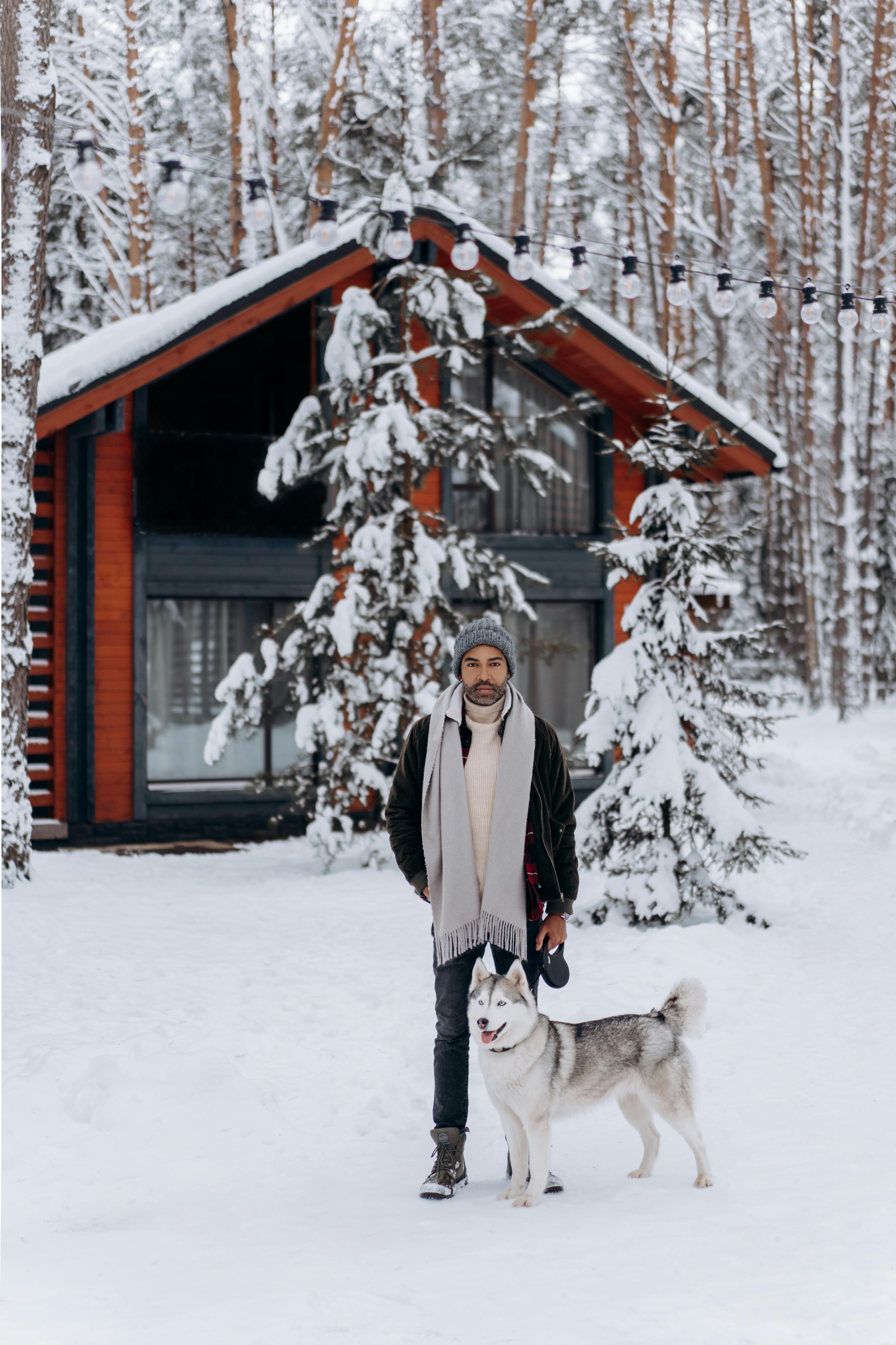 A man stands with a Siberian Husky in front of a snow-covered cabin in a winter forest setting.
