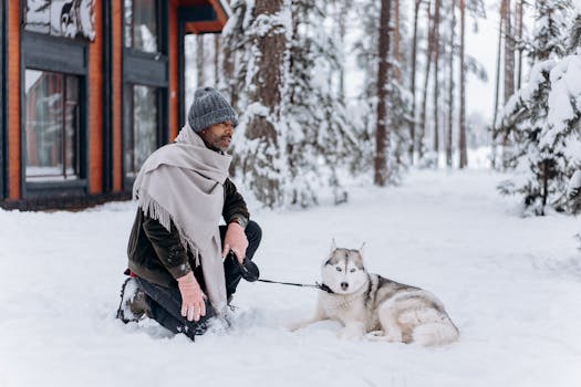 Man kneels beside husky in snow near cabin, enjoying a serene winter forest scene.