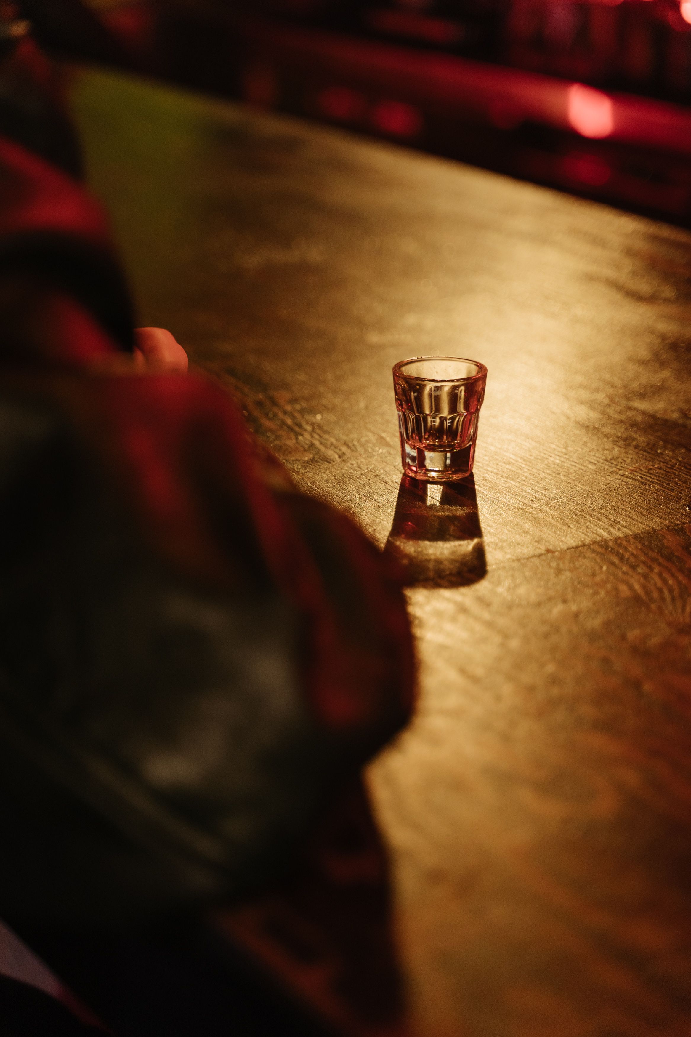 A dimly lit bar counter with a single shot glass casting a shadow.