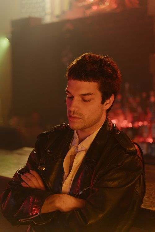 Free Man with arms crossed, wearing a black leather jacket, gazing down in dim lighting. Stock Photo