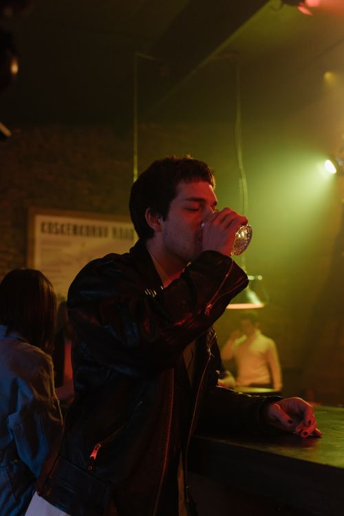Free A young man in a leather jacket enjoys a drink at a dimly lit bar. Stock Photo