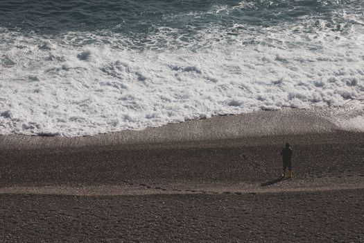 A person stands alone on a rocky beach, watching the waves crash onto the shore.
