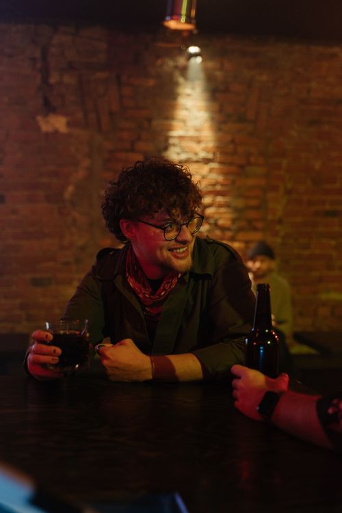 Free Man with curly hair and eyeglasses enjoying a drink and conversation at a rustic bar. Stock Photo