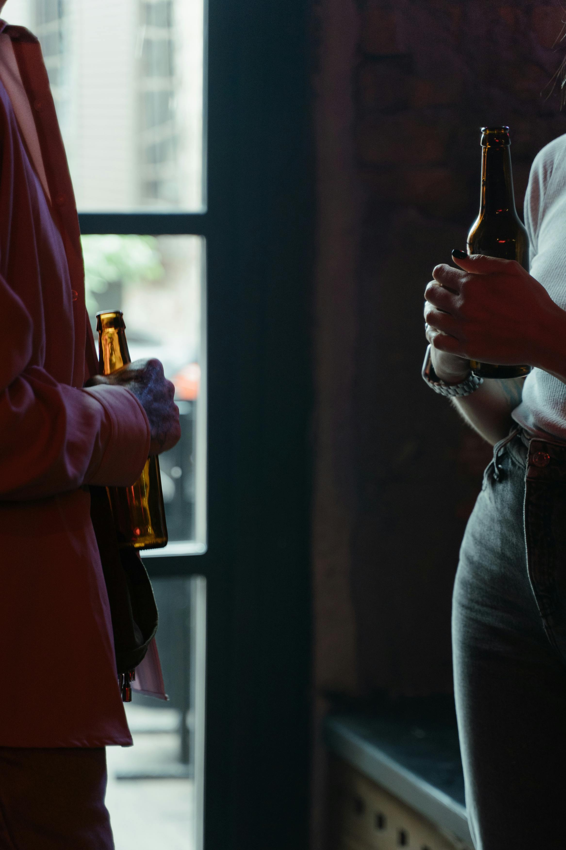 A scene of two people holding beer bottles and talking in a dimly lit bar.