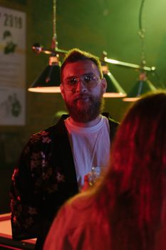 A bearded man with glasses in a dimly lit bar, enjoying a casual evening setting.